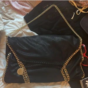 Svart falabella bag från Stella McCartney - Säljer en svart axelväska från Stella McCartney med snygga guldfärgade kedjedetaljer runt kanterna och som axelrem. Väskan har en mjuk, slouchy form och är tillverkad i syntetmaterial. Perfekt för dig som vill ha en statementväska med lyxig känsla. Väskan är i superfint skick och kostar som ny runt 11 000!