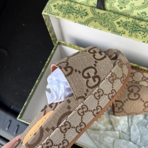 Gucci sandaler med GG-mönster - Snygga sandaler från Gucci med klassiskt GG-mönster i brunt och beige. Sandalerna har bred rem över foten och platt sula med räfflad undersida. Tillverkade i canvas med detaljer i skinn och märkning på innersulan. Perfekta för en trendig sommarlook.
