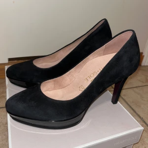 Svarta mockapumps från Tamaris, stl 39 - Snygga svarta pumps från Tamaris Heart & Sole i mocka. Skorna har en plattform framtill och innersula i skinn för extra komfort. Perfekta för dig som vill ha en klassisk och elegant look med en modern twist.