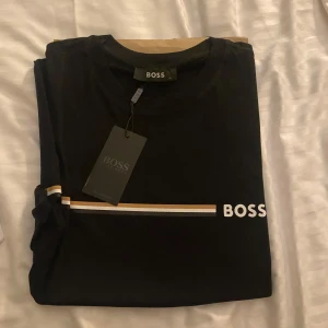 Svart BOSS t-shirt med ränder - Svart t-shirt från BOSS med korta ärmar och rund hals. Framtill finns en vit och beige rand samt BOSS-logga tryckt på bröstet. Tillverkad i mjuk bomull för en skön känsla och clean look. Perfekt för dig som gillar stilrena plagg med en touch av premium.
