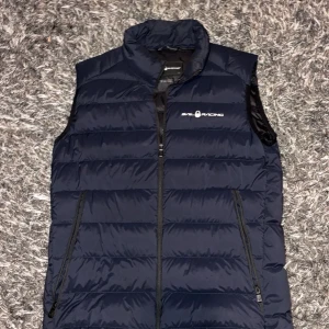 Sailracing Väst - Snygg mörkblå dunväst från Sail Racing med quiltad design och dragkedja framtill. Västen har hög krage och två praktiska fickor med dragkedja. Perfekt för lager-på-lager och sportig stil.