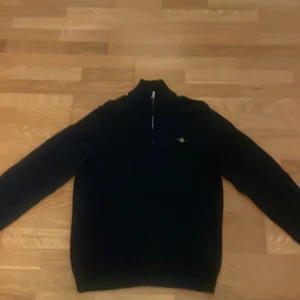 Svart stickad tröja med half zip från Gant - Svart stickad tröja med hög krage och half zip-dragkedja. Tröjan har lång ärm och en liten broderad logga på bröstet. Perfekt för kyligare dagar och enkel att matcha med jeans eller chinos.