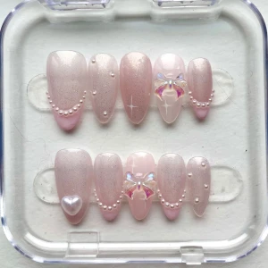 Rosa press-on naglar med pärlor & rosetter - Säljer ett handmålat set med press-on naglar i ljusrosa och skimrande finish. Naglarna är dekorerade med små pärlor, 3D-rosetter i regnbågsskimrande material och ett hjärtformat pärlmotiv. Köp annonsen och skriv din storlek (sticky tabs ingår) ❤️