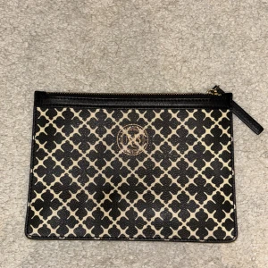 Kuvertväska från By Malene Birger - Snygg kuvertväska från By Malene Birger i svart och beige med ikoniskt grafiskt mönster. Väskan har guldfärgad logga framtill och dragkedja upptill. Tillverkad i tåligt syntetmaterial med beige innerfoder. Perfekt för att bära det viktigaste med stil.