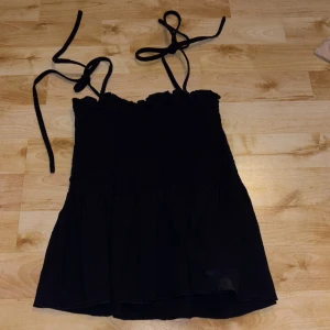 Svart smockad linnetopp från SHEIN - Svart linnetopp från SHEIN i storlek XS. Toppen har smockad överdel, volangkant nedtill och tunna axelband som knyts på axlarna. Perfekt för varma dagar och enkel att styla med jeans eller kjol.