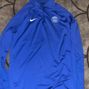  PSG  träningströja Nike - Säljer en blå långärmad fotbollströja från Nike med PSG-logga på bröstet. Tröjan är i Dri-Fit-material och har halv dragkedja framtill. Slim fit-modell med diskret mönster på ärmarna. Perfekt för träning eller match.