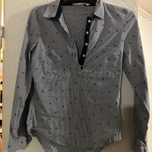 Randig henley-skjorta från Zara Basic - Snygg randig henley-skjorta från Zara Basic i blått och vitt med små mörkblå broderade detaljer. Skjortan har lång ärm, knappar framtill och två bröstfickor. Perfekt för en chill och stilren look.