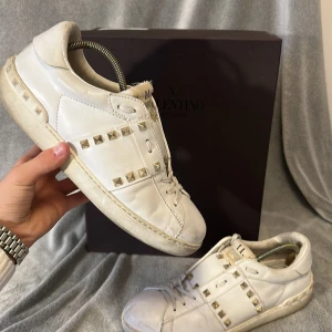 Valentino Rockstud vita sneakers - Snygga vita Valentino Rockstud sneakers i skinn med ikoniska guldiga nitar längs sidorna. Klassisk låg modell med snörning och rund tå. Perfekta för dig som vill ha en lyxig och edgy touch till din outfit. Box medföljer ej