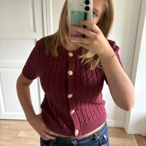 Vinröd kabelstickad kofta med guldknappar - Säljer en vinröd kabelstickad kofta med korta ärmar och snygga guldknappar framtill. Tröjan har ribbade kanter och en croppad passform och knappt använd.