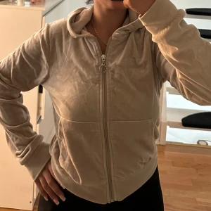 Beige velour hoodie med dragkedja - Mysig beige hoodie i mjuk velour med dragkedja framtill och huva. Tröjan har långa ärmar, fickor på sidorna och en snygg ringdetalj på dragkedjan. Perfekt för chill dagar eller när du vill vara extra comfy. Snöret till luvan är dock borttappad.