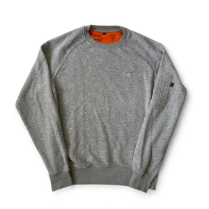 Grå sweatshirt från Alpha Industries | S - Säljer en grå sweatshirt från Alpha Industries med klassisk rund hals och orange insida. Tröjan har en liten logotyp på bröstet och en ärmficka med dragkedja och röd 'Remove Before Flight'-tag. Som går att ta av! 