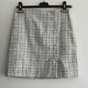 Vit och svart rutig kortkjol med knappar - Snygg kortkjol i vit och svart rutigt tyg med dekorativa knappar framtill. Kjolen har en rak passform och är fodrad på insidan för extra komfort. Perfekt för dig som gillar en stilren och trendig look.