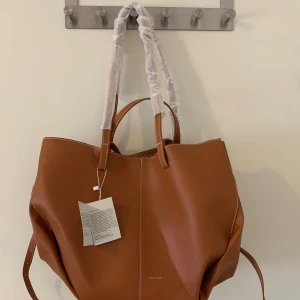 Polène brun axelväska i skinn - Snygg och rymlig brun axelväska från Polène i slätt skinn med vita kontrastsömmar. Väskan har både korta och långa handtag samt en matchande innerpouch. Perfekt för dig som vill ha en stilren och trendig väska med plats för allt du behöver.