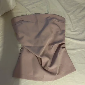 Ljusrosa axelbandslös tubtopp - Säljer en stilren ljusrosa tubtopp med bandeau-modell. Toppen är axelbandslös och har en slät, enkel design med lätt utsvängd form nedtill. Tillverkad i ett mjukt, slätt syntetmaterial med insydd resår upptill för att sitta på plats.