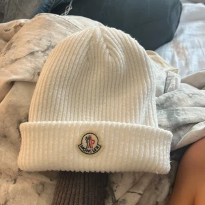 Vit ribbad Moncler mössa - Snygg vit ribbstickad mössa från Moncler (det är en replika)med klassisk logotyp framtill. Mössan har uppvikt kant och är tillverkad i mjukt stickat material som håller dig varm och stilren under kalla dagar.