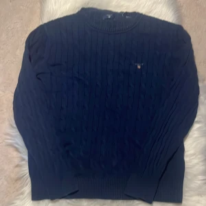 Marinblå kabelstickad tröja från Gant - Kabelstickad marinblå tröja från Gant med broderad logga på bröstet. Klassisk rund halsringning och ribbade muddar. Perfekt för dig som gillar preppy stil och vill ha något snyggt och tidlöst i garderoben.