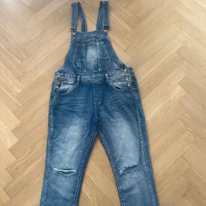 Blå denim hängselbyxor med slitningar - Snygga blå denim hängselbyxor med justerbara axelband och flera fickor fram och bak. Byxorna har slitna detaljer vid knäna och är väldigt bekväma . Perfekta för en avslappnad streetstil. Storleken är L, men de kan även passa M beroende på hur man vill att de ska sitta.  De är aldrig använda och i perfekt skick!