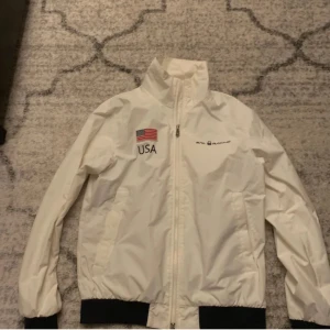 Vit vindjacka Sail Racing USA - Vit vindjacka från Sail Racing med USA-flagga och texten 'USA' fram samt 'UNITED STATES' och flagga bak. Jackan har svarta muddar, hög krage och dragkedja framtill. Perfekt för dig som gillar sportig och clean stil.