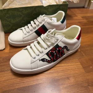 Gucci Ace sneakers med orm brodyr - Säljer ett par vita Gucci Ace sneakers i skinn med ikonisk röd och svart orm brodyr på sidan, gröna och röda detaljer samt röd häl i ormmönstrat material. Klassisk rund tå, snörning och platt sula. Riktigt snygga och trendiga sneakers för dig som vill sticka ut.