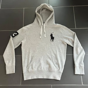 Grå hoodie från Polo Ralph Lauren - Snygg ljusgrå hoodie från Polo Ralph Lauren med stor svart logga på bröstet och vit snörning i huvan. Tröjan har en känguruficka framtill och en svart siffra på ärmen. Tillverkad i mjuk bomulls- och polyestermix. Passar S-M. Pris kan såklart diskuteras 