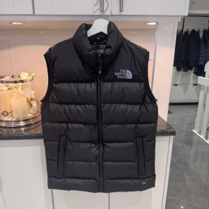 Svart dunväst från The North Face - Säljer en svart dunväst från The North Face med hög krage och dragkedja framtill. Västen är quiltad och har två fickor på sidorna. Perfekt för lager-på-lager och har en clean look med logga på bröstet och ryggen. Modellen är klassisk och passar till många stilar. Helt oanvänd. Nypris 2000kr