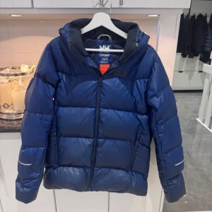 Blå Helly Hansen pufferjacka 176/16 - Snygg blå pufferjacka från Helly Hansen med huva och dragkedja framtill. Jackan har en quiltad look, två sidofickor med dragkedja och reflekterande detaljer på ärmarna. Perfekt för kalla dagar när du vill hålla värmen och ändå se cool ut. Helt oanvänd!