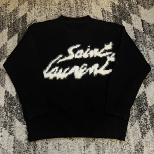 Svart stickad Saint Laurent tröja - Svart stickad tröja från Saint Laurent med vit, fluffig logga framtill. Tröjan har rund halsringning, ribbade muddar och en loose passform. Perfekt för dig som vill ha en statement piece med lyxig känsla.