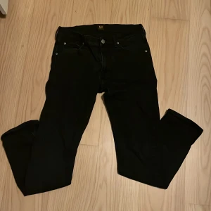 Svarta jeans från Lee - Säljer ett par svarta jeans från Lee med klassisk femficksdesign. Jeansen har bälteshällor, knapp- och dragkedjestängning samt Lee-logga bak i midjan. Materialet är jeans och färgen är helsvart, vilket gör dem enkla att matcha.