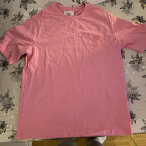 Rosa t-shirt från Ami med hjärtdetalj - Snygg rosa t-shirt från Ami Alexandre Mattiussi med broderad hjärtdetalj på bröstet. Klassisk passform, rund hals och korta ärmar. Tillverkad i mjuk bomull som känns skön mot huden. Perfekt för dig som gillar stilrena plagg med en twist.
