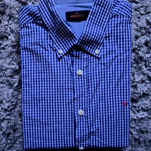 Blårutig skjorta från Morris - Snygg blårutig skjorta från Morris med klassisk krage och vita knappar. Skjortan har ett broderat rosa emblem på bröstet och är tillverkad i bomull. Perfekt för dig som gillar stilrena och tidlösa plagg.