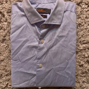 Ljusblå skjorta från Morris - Snygg ljusblå skjorta från Morris med klassisk krage och broderad logga på bröstet. Skjortan har knappar framtill och är tillverkad i mjuk bomull. Perfekt för dig som gillar stilrena och enkla plagg med en clean look.