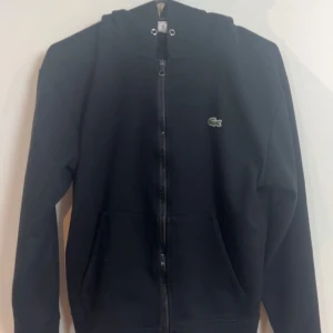 Svart hoodie från Lacoste med dragkedja - Svart hoodie från Lacoste med klassisk krokodillogga på bröstet. Tröjan har huva, hel dragkedja framtill och två fickor. Perfekt för chill dagar och enkel att matcha med jeans eller joggers.