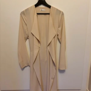Beige trenchcoat med knytband - Stilren beige trenchcoat från C i storlek xs Jackan har långa ärmar, öppen front med snyggt fall och knytband i midjan för en schysst siluett. Perfekt för dig som gillar minimalistisk och clean stil.