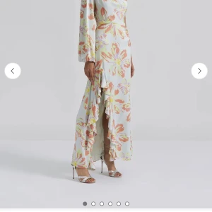 Långklänning By Malina med volang - Säljer en superfin långklänning med stora blommor i ljusa färger som vitt, orange, gul och lila. Klänningen har långärmade ärmar, v-ringning och en snygg volangdetalj längs slitsen framtill. Perfekt för dig som gillar romantiska och färgglada plagg.