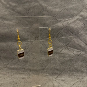 Örhängen med kaffemugg-hängen - Unika örhängen med små kaffemuggar som hängsmycke. Muggarna är vita med brunt band där det står 'coffee' och detaljer i guld. Perfekta för dig som älskar kaffe och vill ha något lekfullt i din stil.