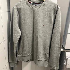 Grå sweatshirt från Tommy Hilfiger - En stilren grå sweatshirt från Tommy Hilfiger med rund halsringning och diskret logga broderad på bröstet. Tröjan har långa ärmar och ribbade muddar vid ärmslut och nederkant. Perfekt för en avslappnad och snygg look.