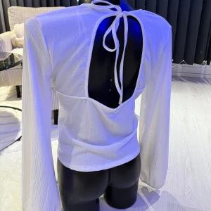 Vit långärmad blus med öppen rygg - Säljer en vit blus med lång ärm och snygg v-ringning framtill. Toppen har öppen rygg med knytband i nacken och lätt strukturerat tyg. Perfekt för dig som vill ha en stilren men ändå unik look. Passar som oversize modell.