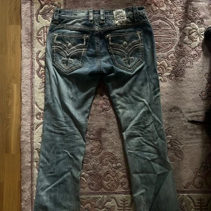 Rock Revival jeans med broderade fickor - Snygga blå jeans från Rock Revival med markerade kontrastsömmar och broderade bakfickor med dekorativa detaljer. Märkespatch i vitt på baksidan och coola nitar på fickorna. Ska dubbelkolla storleken, skicka gärna DM om du är intresserad