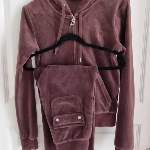 Brun velour hoodie Juicy Couture - Mysig brun hoodie och byxor från Juicy Couture i mjuk velour med dragkedja framtill, snörning i huvan och broderad logga på bröstet. Långa ärmar och ribbade muddar. Perfekt för chill dagar eller när du vill vara extra comfy. Xs på tröjan och xxs på byxorna 