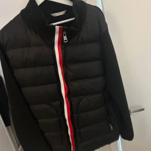 Svart Moncler pufferjacka med röd/vit dragkedja - Snygg svart pufferjacka från Moncler med stickade ärmar och coolt Moncler-märke på armen. Jackan har en dragkedja framtill med röd och vit detalj som ger en sportig vibe. Perfekt för dig som vill ha en stilren och trendig look under kyliga dagar. Xl passar L 185-190 cm