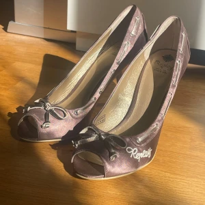 Bruna Replay pumps med rosett, strl 39 - Snygga bruna pumps från Replay i satinliknande material med öppen tå och rosett framtill. Skorna har broderad Replay-logga på sidan och dekorativa snörningsdetaljer längs kanten. Klackarna är medelhöga och har en trälook. Perfekta för dig som vill sticka ut lite extra.