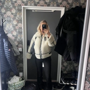 Vit teddyjacka med svarta detaljer - Säljer en fluffig vit teddyjacka med svarta kantdetaljer och dragkedja framtill. Jackan har stor krage, markerade sömmar och en cool, oversized passform. Perfekt för dig som vill ha en trendig och mysig look.