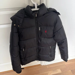 Ralph Lauren jacka - Säljer en svart pufferjacka från Ralph Lauren med röd logga på bröstet. Jackan har huva, dragkedjor på fickorna och en hög krage. Perfekt för kalla dagar och riktigt snygg till streetwear-stilen. Pris kan diskuteras vid snabb affär 