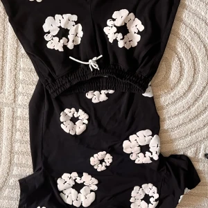 Svarta shorts från Sporty & Rich S - Svarta shorts från the denim tears med vita blommiga tryck över hela plagget. De har elastisk midja med vit snörning och är tillverkade i mjuk bomull. Perfekta för en chill och trendig sommarlook. Man får både tröjan och shortsen för bara 1300 kr