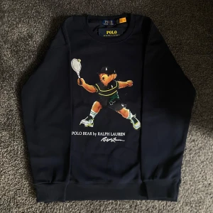 Mörkblå sweatshirt Polo Bear Ralph Lauren - Snygg mörkblå sweatshirt från Ralph Lauren med Polo Bear-tryck där björnen spelar tennis. Tröjan har rund hals, långa ärmar och ribbade muddar. Perfekt för dig som gillar sportiga och ikoniska prints. Materialet är mjuk bomull som känns skönt mot huden. Nypris 