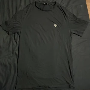 Svart t-shirt från Emporio Armani EA7 - Svart t-shirt från Emporio Armani EA7 med liten logga på bröstet. Klassisk rund hals och korta ärmar. Tillverkad i mjukt material som känns skönt mot huden. Perfekt för en clean och stilren look.