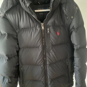 Svart pufferjacka från Polo Ralph Lauren - Svart pufferjacka från Polo Ralph Lauren med röd broderad logga på bröstet. Jackan har huva, flera dragkedjeförsedda fickor och kraftig vaddering som håller dig varm. Klassisk och stilren design med raka linjer och hög krage.