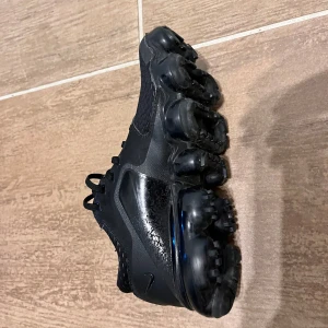 Nike Vapormax svarta sneakers - Svarta Nike Vapormax sneakers med unik bubblig sula och snörning. Ovandelen är i mesh och syntet för en sportig look. Swoosh-logga på sidan och diskret design gör dem riktigt cleana. Perfekta för dig som gillar streetstyle och vill ha något som sticker ut.