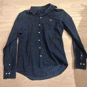 Mörkblå mönstrad skjorta från Gant - Snygg mörkblå skjorta från Gant med små vita mönster över hela tyget. Klassisk krage, vita knappar och lång ärm. Broderad Gant-logga på bröstet. Perfekt för dig som gillar stilrena detaljer och tidlös design. Nästan helt oanvänd.