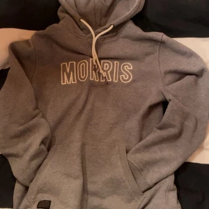 Grå morris hoodie - Grå hoodie från Morris med vit snörning i huvan och stort MORRIS-tryck på bröstet. Perfekt för chill dagar eller när du vill ha en enkel och snygg look. Hoodien är använd men ändå i bra skick!